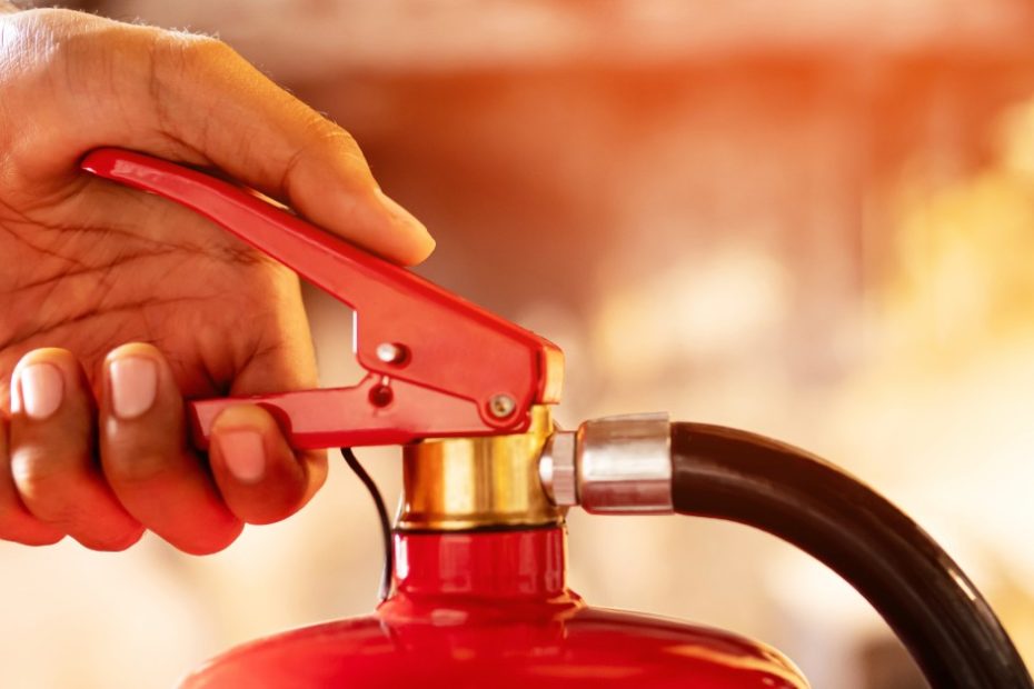 A hand prepares to press down on the trigger of a fire extinguisher as flames roar in the background.