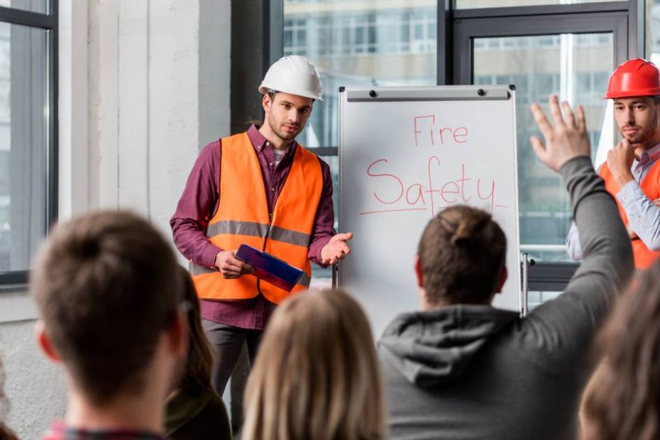 Two men in safety gear stand before a whiteboard reading "Fire Safety" in red letters and answer questions from the class they're teaching.