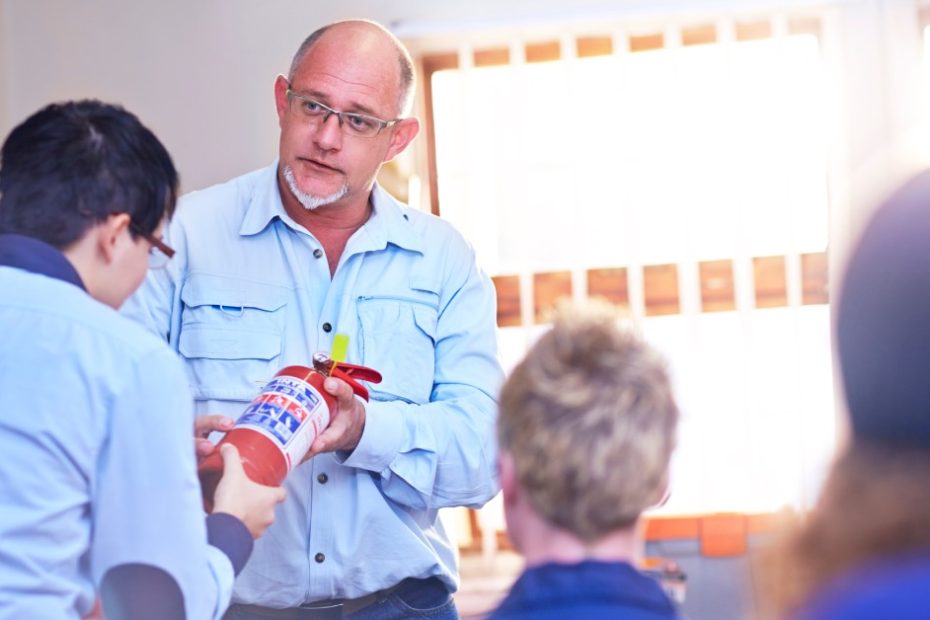 An instructor showing his students a fire extinguisher. One student studies the extinguisher closely, the others look on from afar.