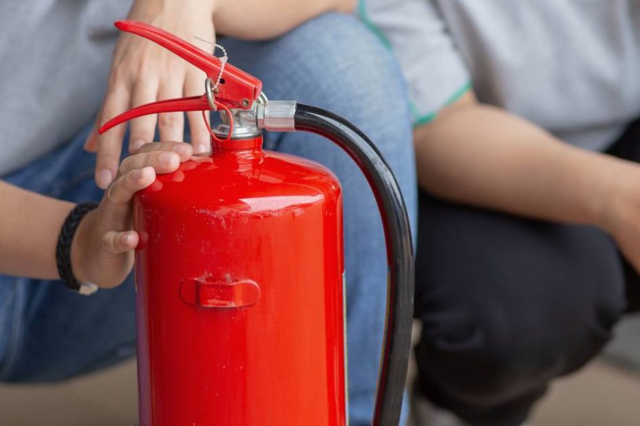 Two people dressed in gray shirts kneeling behind a fire extinguisher. One of them rests a hand on the extinguisher.