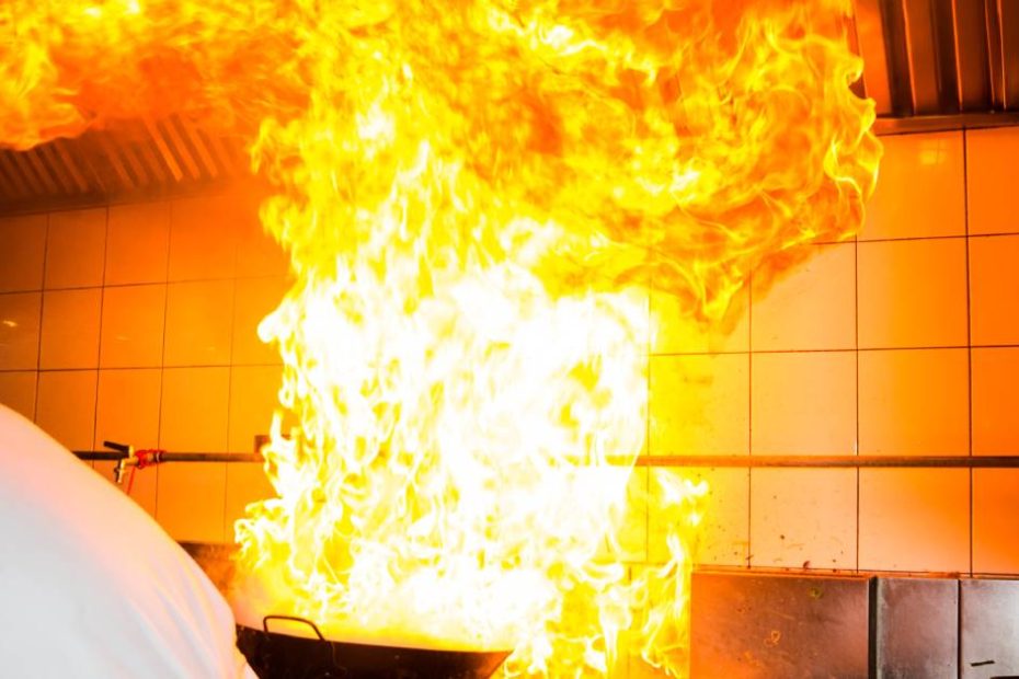 Flames pouring from a wok on a stovetop as a chef moves away. The flames billow all the way up toward the ceiling.