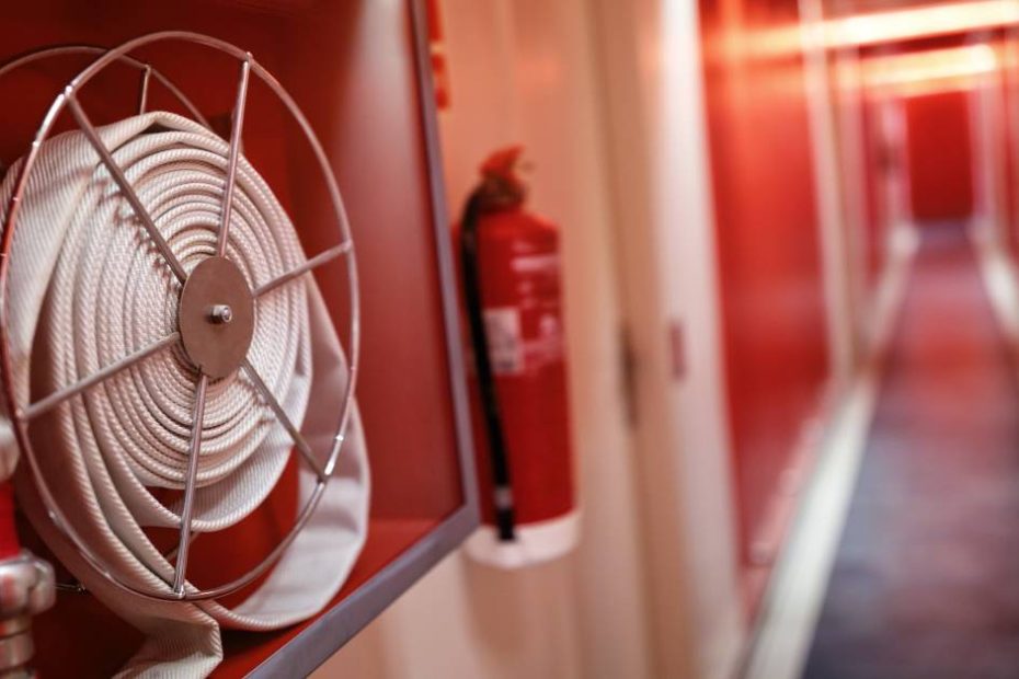 A fire extinguisher and a fire hose mounted on the wall of a narrow corridor, which is painted a shade of red.