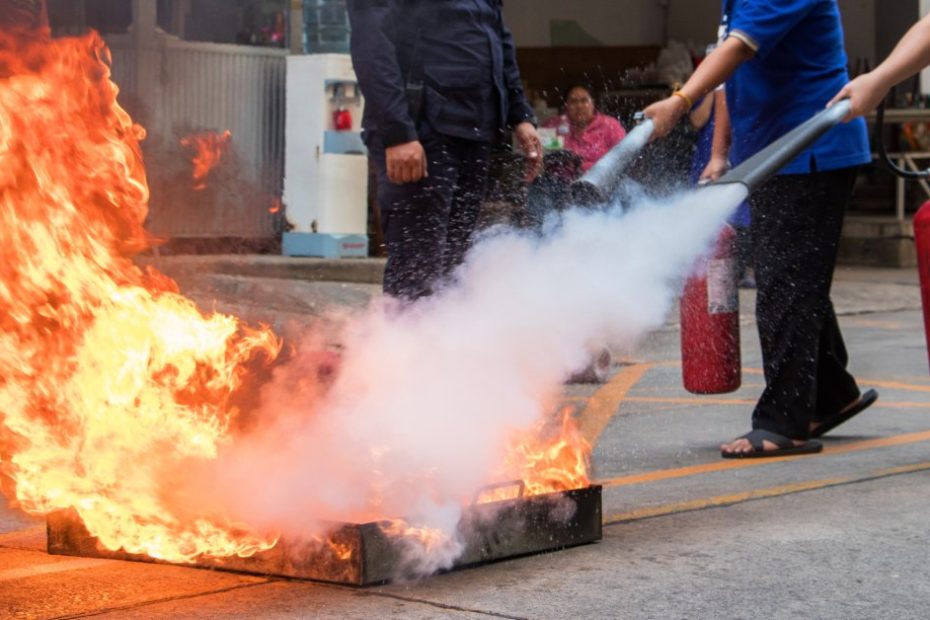 Two employees in blue polo shirts using fire extinguishers on a large fire in a metal box for a lesson in a parking lot.