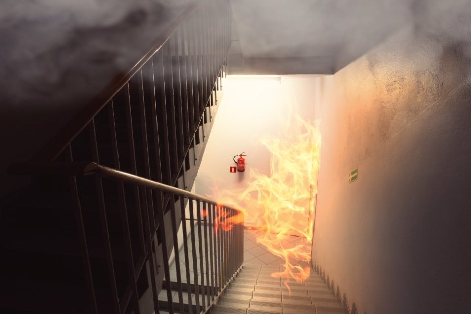 A fire is burning in the stairwell of a building. At the base of a flight of stairs, flames blaze near a fire extinguisher.